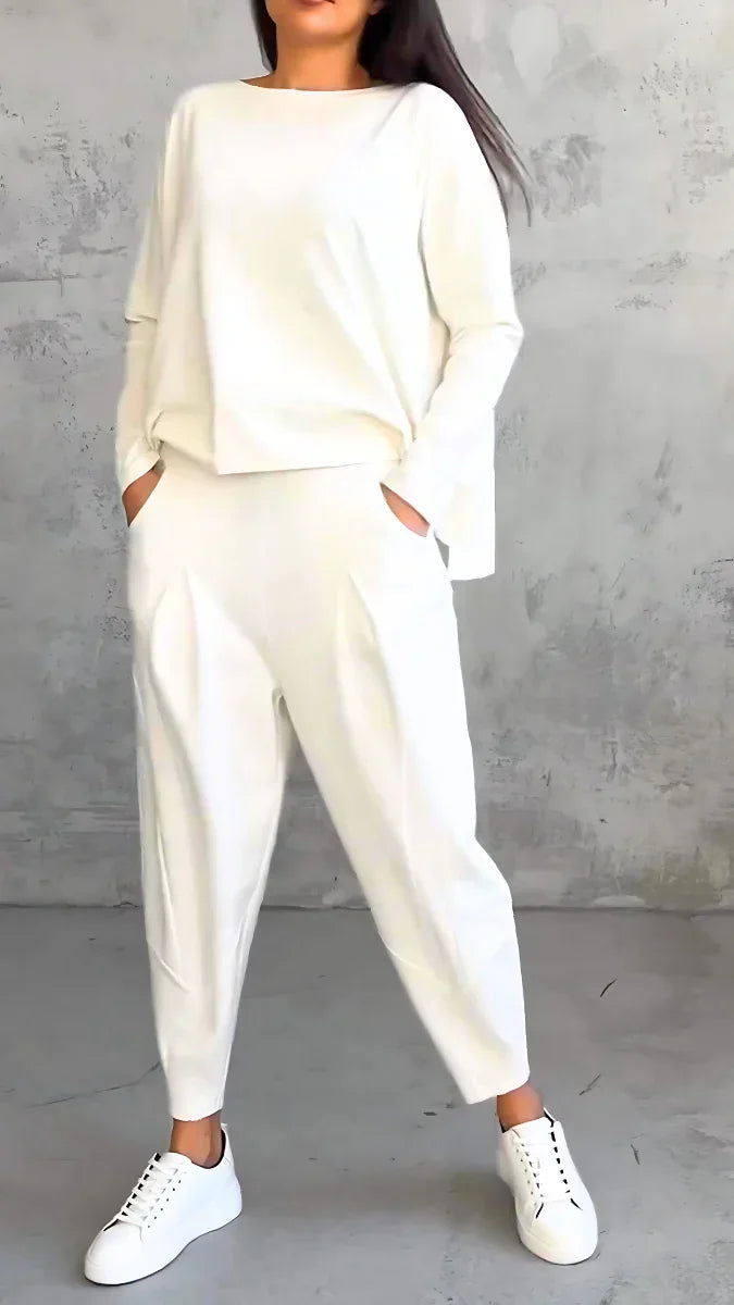 Frau in weißem Oversize-Pullover und Hose, weiße Sneaker, grauer Hintergrund.