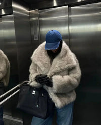 Person in beiger Kunstpelzjacke, blauer Jeans und blauer Kappe in Aufzug mit schwarzer Tasche.