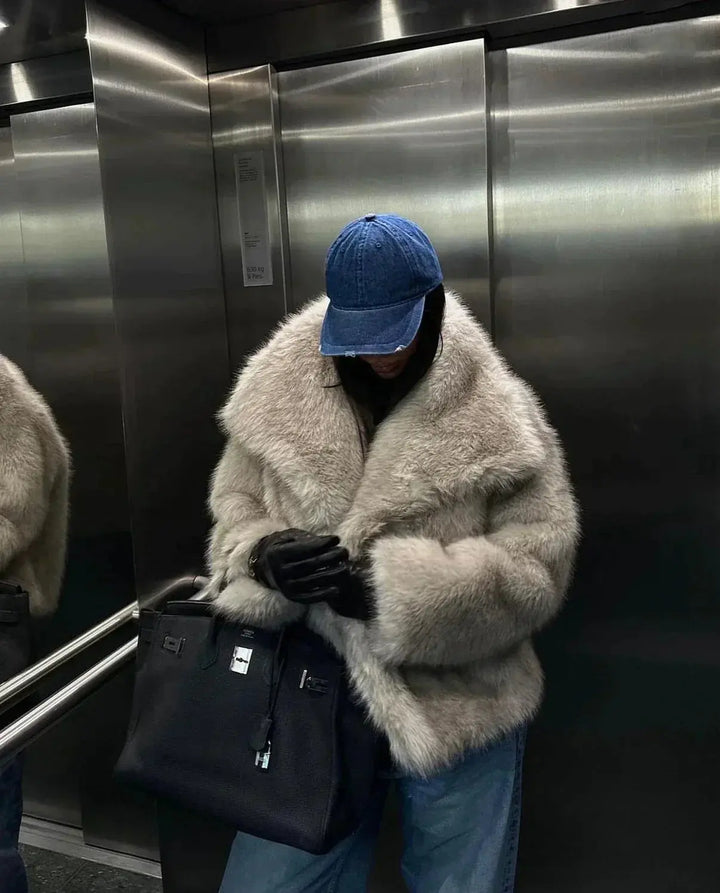 Person in beiger Kunstpelzjacke, blauer Jeans und blauer Kappe in Aufzug mit schwarzer Tasche.