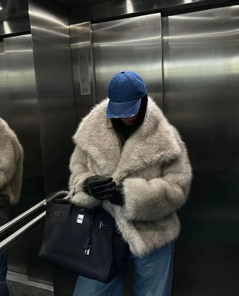 Person in beiger Kunstpelzjacke, blauer Jeans und blauer Kappe in Aufzug mit schwarzer Tasche.