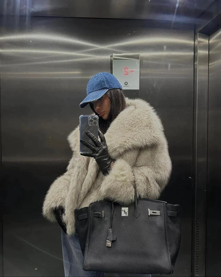 Frau im Aufzug mit Pelzmantel, blauer Kappe, schwarzer Lederhandtasche, Smartphone in der Hand.