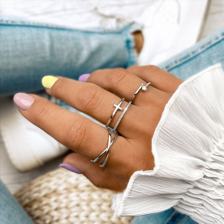 Frau mit silbernen Ringen, Kreuzdesign, pastellfarbene Nägel, weiße Bluse, Jeans.