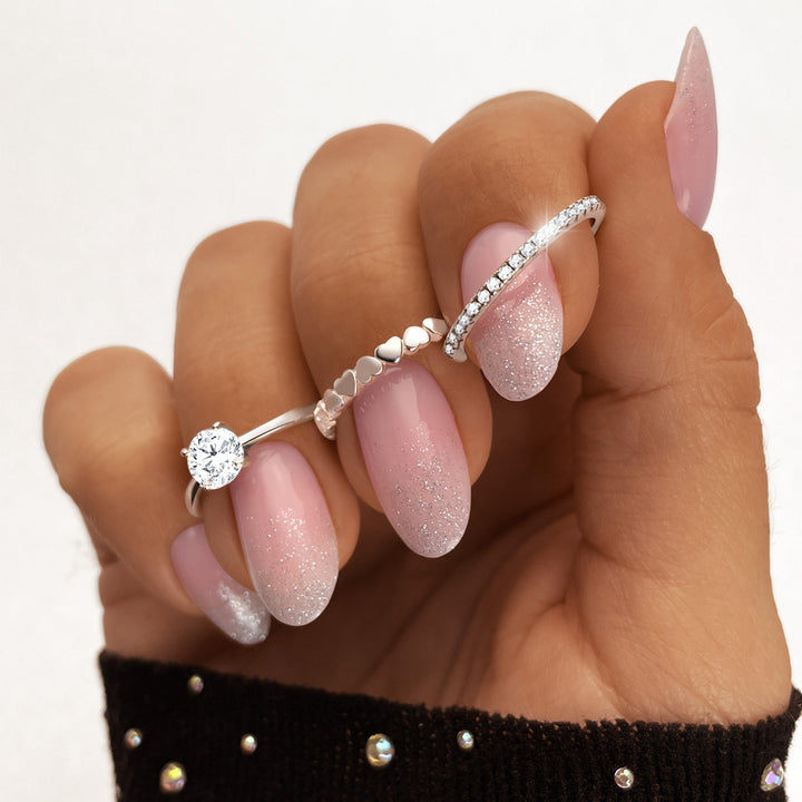 Drei silberne Ringe mit Herz- und Diamantdesign auf glitzernd lackierten Fingernägeln.