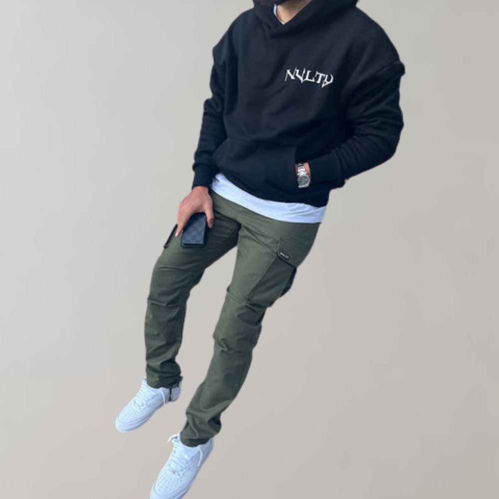 Mann in schwarzem Hoodie, olivgrüner Cargohose, weißen Sneakers, hält Smartphone.