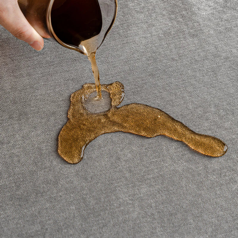 Hand gießt Flüssigkeit auf grauen, wasserabweisenden Stoffbezug, Fleckenschutz, Textilpflege.