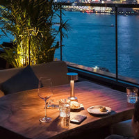Terrasse bei Nacht mit Holztisch, Weinglas, Kerzenlicht, Blick auf beleuchtete Stadt und Fluss.