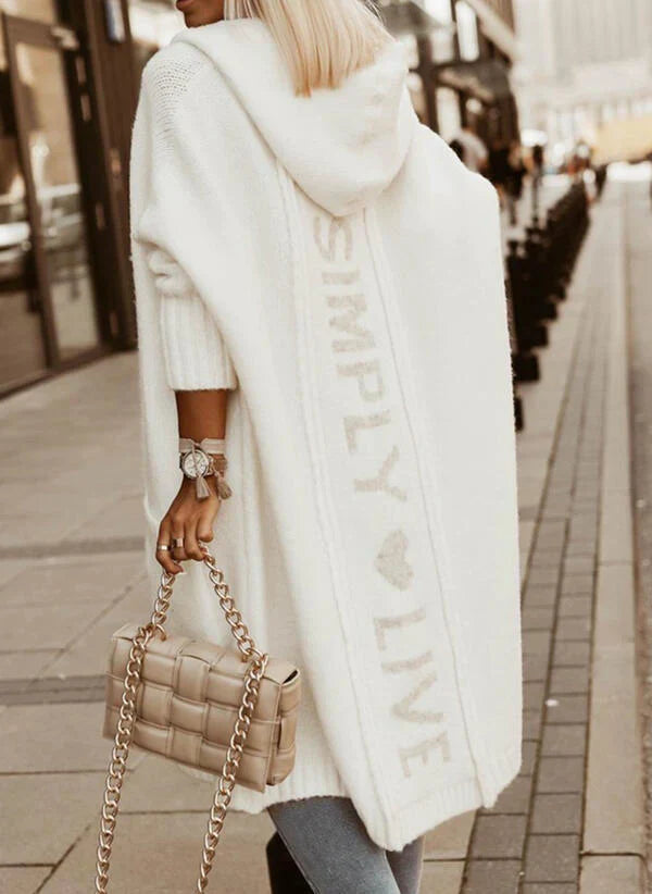 Frau in weißem Strickmantel mit Kapuze und Schriftzug, beige gesteppte Handtasche, Stadtstraße.