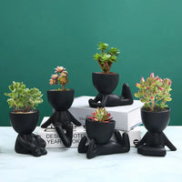 Schwarze Keramik-Blumentöpfe in menschlicher Form mit Sukkulenten auf grünem Hintergrund.