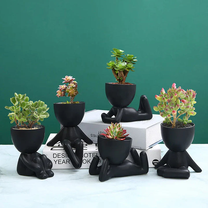 Schwarze Keramik-Blumentöpfe in menschlicher Form mit Sukkulenten auf grünem Hintergrund.