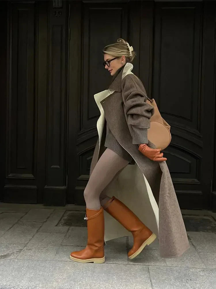 Frau in braunem Mantel, beigen Stiefeln und orangefarbenen Handschuhen vor dunkler Tür.