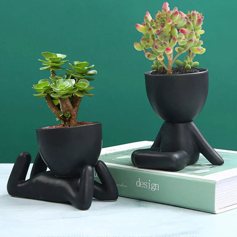 Zwei schwarze Keramik-Blumentöpfe in menschlicher Form mit Sukkulenten auf grünem Hintergrund.