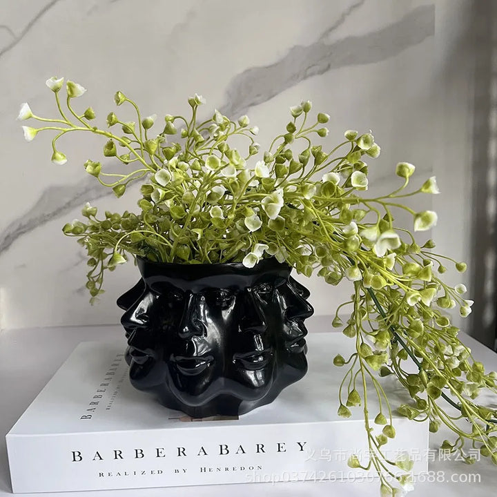 Schwarzer Keramik-Blumentopf mit Gesichtern, gefüllt mit weißen Blumen, auf Buch platziert.