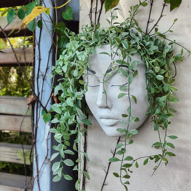 Weiße Gesichtsmaske aus Beton mit hängenden grünen Pflanzen, Wanddekoration im Garten.