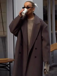 Frau in braunem Wollmantel trinkt Kaffee, trägt Sonnenbrille, modisch, urbaner Stil.