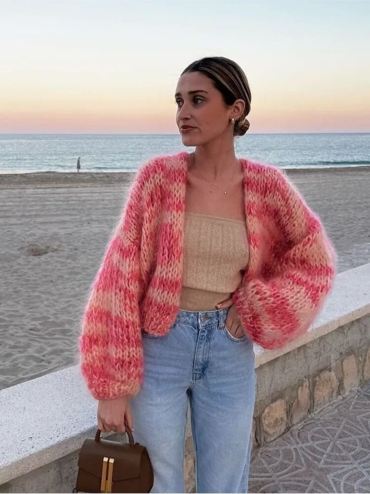Frau am Strand in rosa Strickjacke, beige Top, Jeans; Sonnenuntergang im Hintergrund.