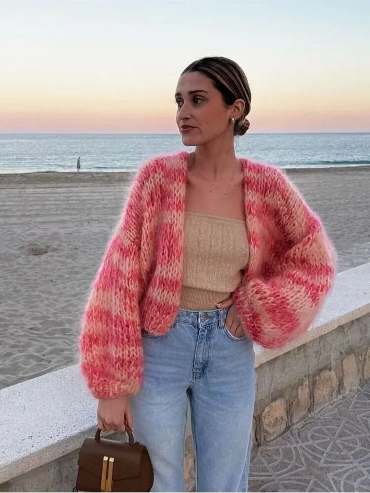 Frau am Strand in rosa Strickjacke, beige Top, Jeans; Sonnenuntergang im Hintergrund.