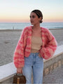 Frau am Strand in rosa Strickjacke, beige Top, Jeans; Sonnenuntergang im Hintergrund.