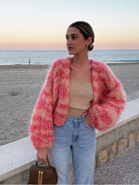 Frau am Strand in rosa Strickjacke, beige Top, Jeans, Sonnenuntergang im Hintergrund.