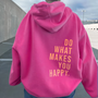 Frau in pinkem Hoodie mit gelbem Schriftzug "Do What Makes You Happy" auf Parkplatz.