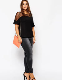 Frau in schwarzer Bluse und grauer Jeans, hält orangefarbene Clutch, trägt schwarze High Heels.