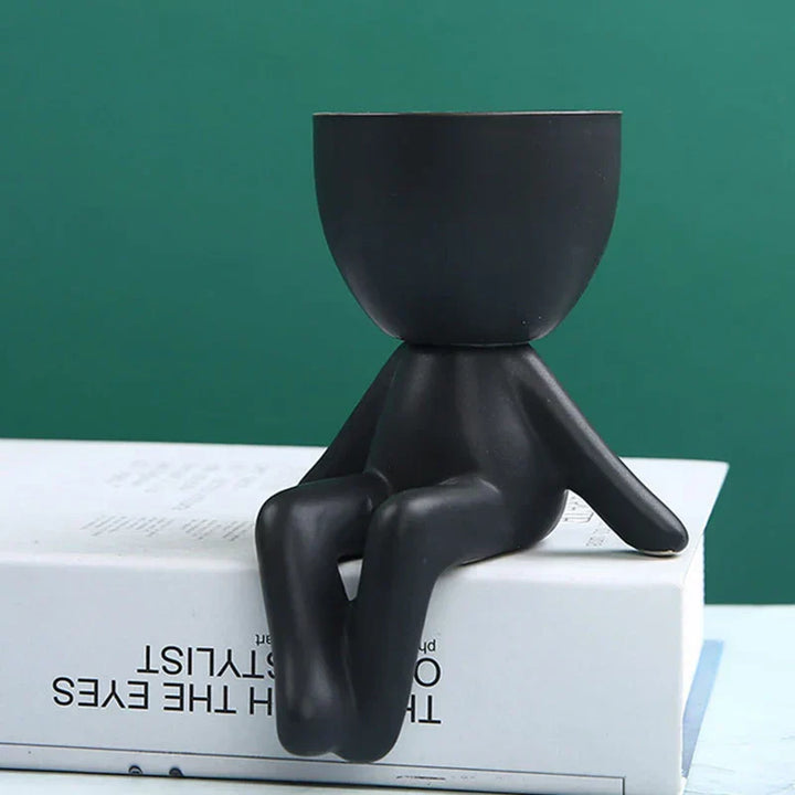 Schwarzer Keramik-Blumentopf in Form einer sitzenden Figur auf Buch, modernes Design.