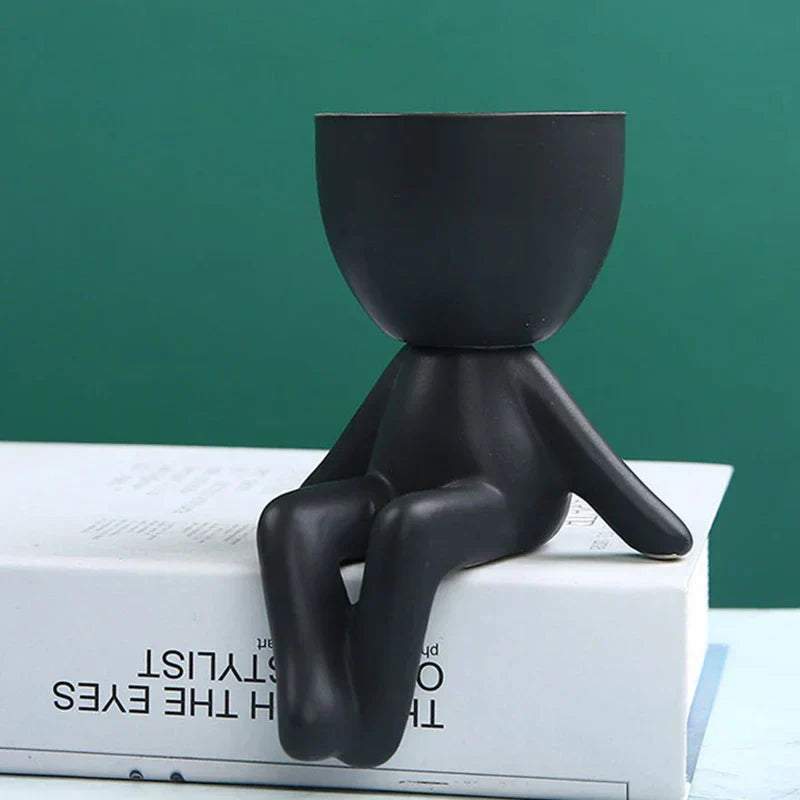 Schwarzer Keramik-Blumentopf in Form einer sitzenden Figur auf Buch, modernes Design.