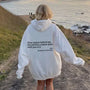 Weiße Oversize-Hoodie mit schwarzem Text, Frau am Küstenweg, lässige Mode, Naturkulisse.