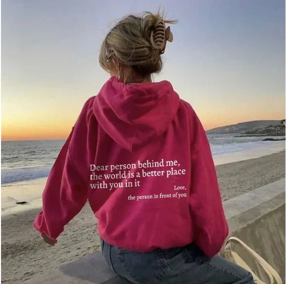 Frau am Strand in pinkem Hoodie mit motivierendem Zitat, Sonnenuntergang im Hintergrund.