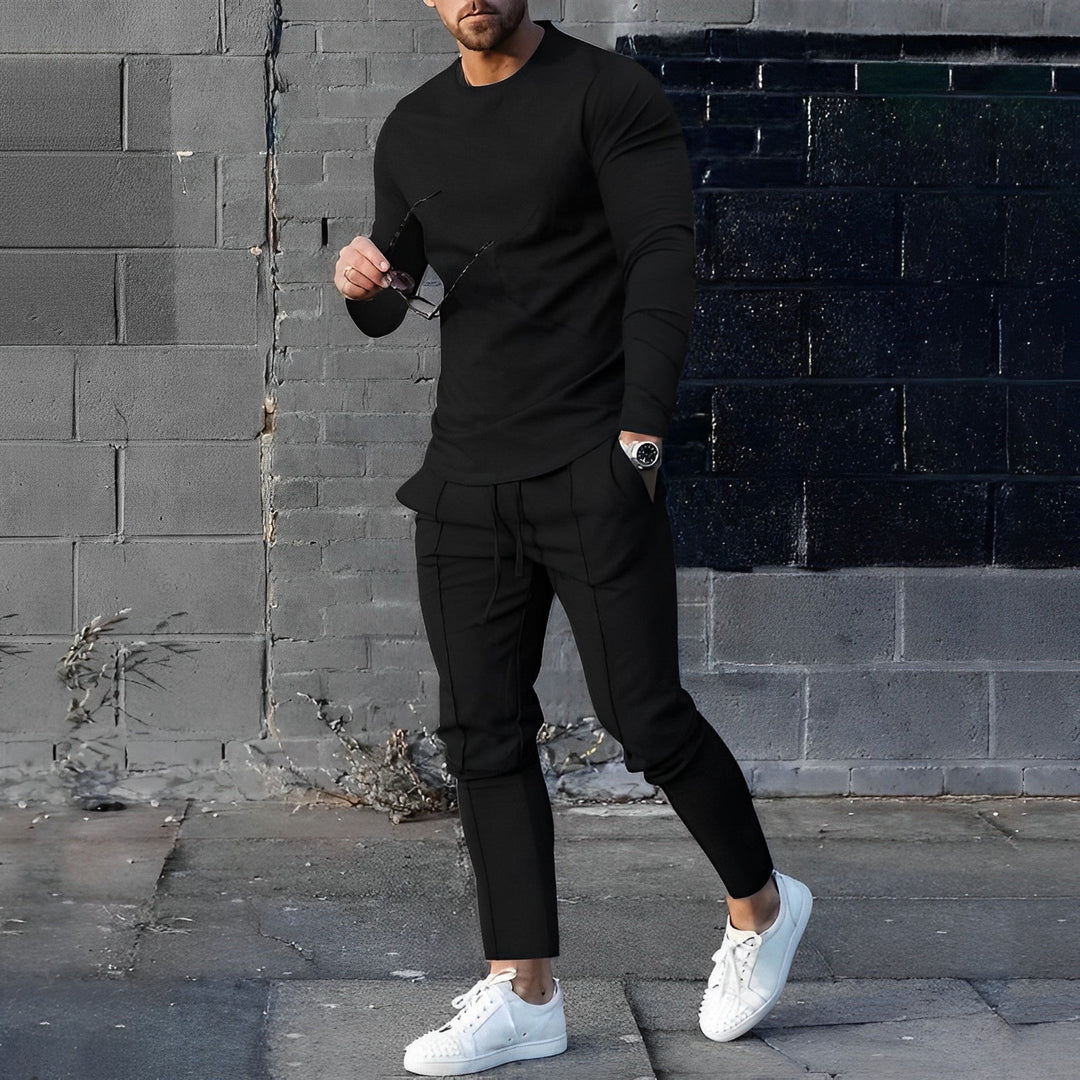 Mann in schwarzem Langarmshirt und Hose, weiße Sneaker, urbane Mode, graue Ziegelwand.