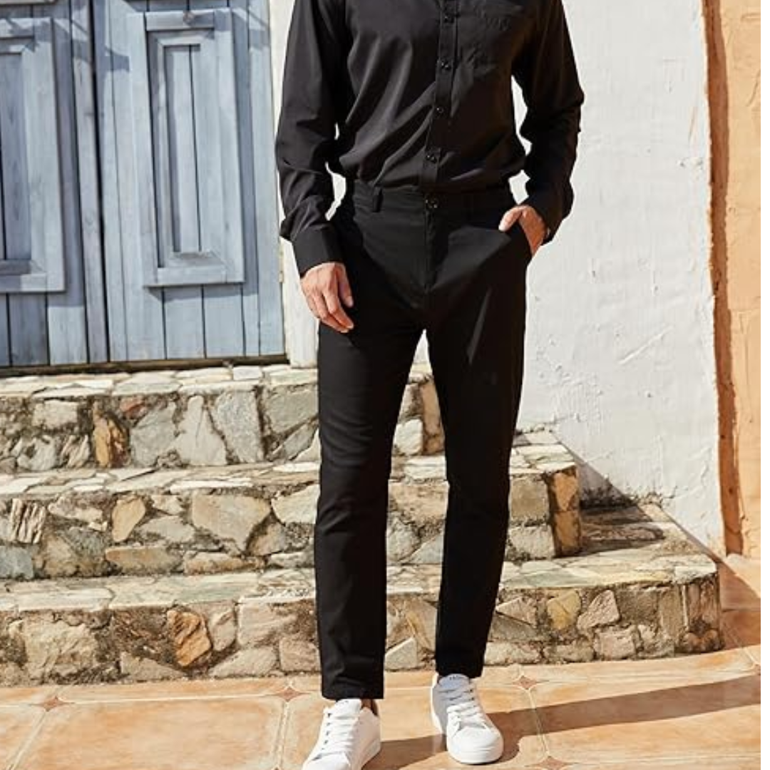 Mann in schwarzem Hemd und Hose, weiße Sneaker, vor blauer Holztür und Steinmauer.