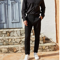 Mann in schwarzem Hemd und Hose, weiße Sneaker, vor blauer Holztür und Steinmauer.