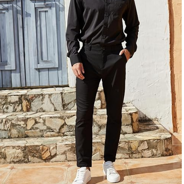 Mann in schwarzem Hemd und Hose, weiße Sneaker, vor blauer Holztür und Steinmauer.