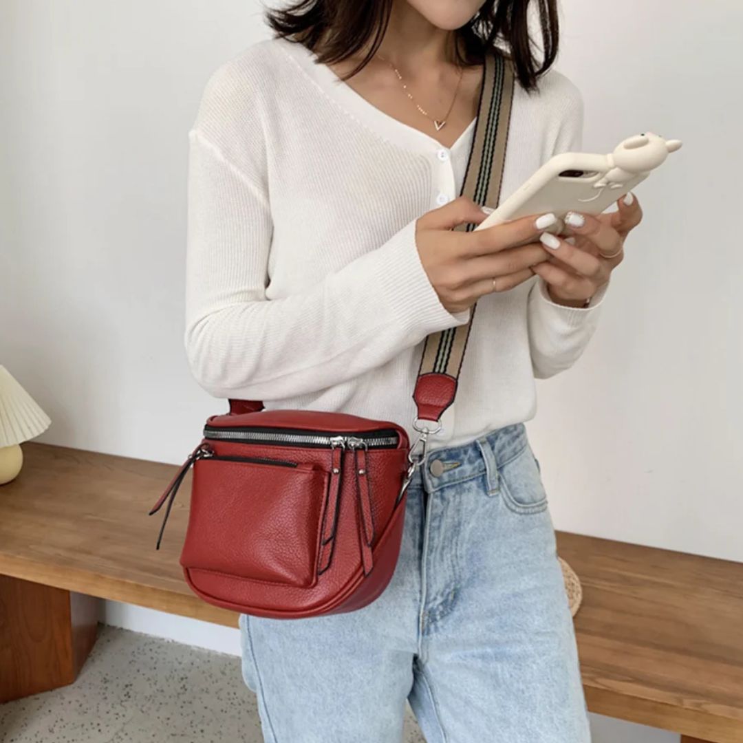 Frau mit roter Leder-Umhängetasche, weißem Pullover, Jeans, Smartphone in modernem Raum.