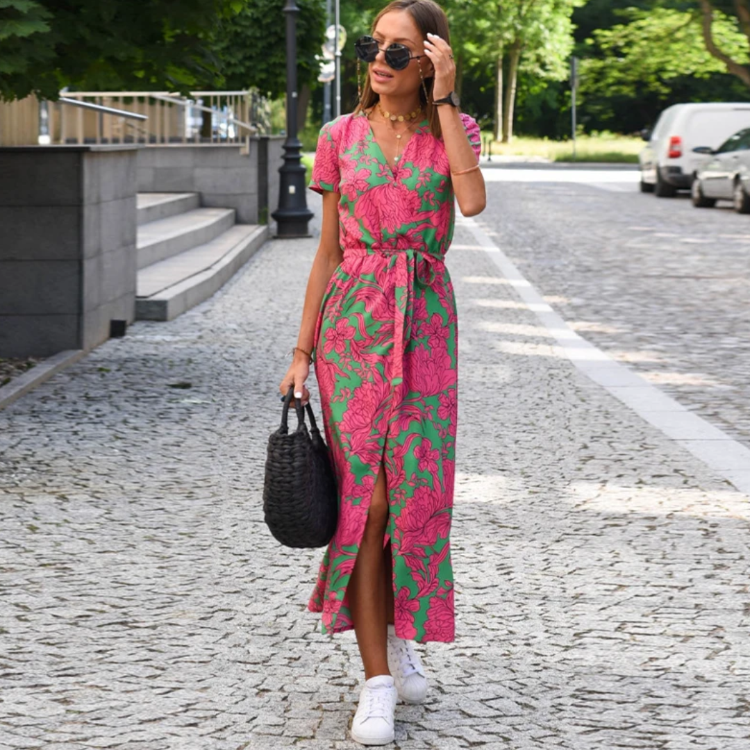 Frau in rosa-grünem Sommerkleid mit Blumenmuster, trägt schwarze Tasche, steht auf gepflasterter Straße.
