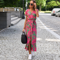 Frau in rosa-grünem Sommerkleid mit Blumenmuster, trägt schwarze Tasche, steht auf gepflasterter Straße.