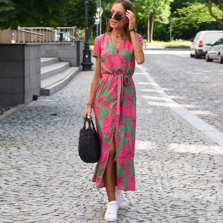 Frau in rosa-grünem Sommerkleid mit Blumenmuster, trägt schwarze Tasche, steht auf gepflasterter Straße.