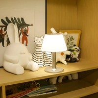 Moderne Tischlampe mit weißem Schirm auf Holzregal, umgeben von Dekofiguren und Büchern.