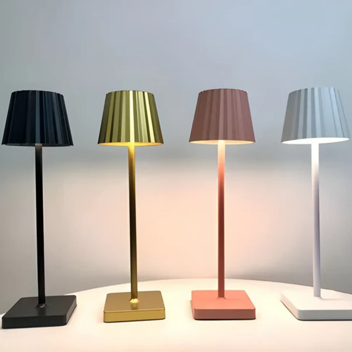 Vier moderne Tischlampen in Schwarz, Gold, Rosa und Weiß auf einem Tisch, minimalistisches Design.