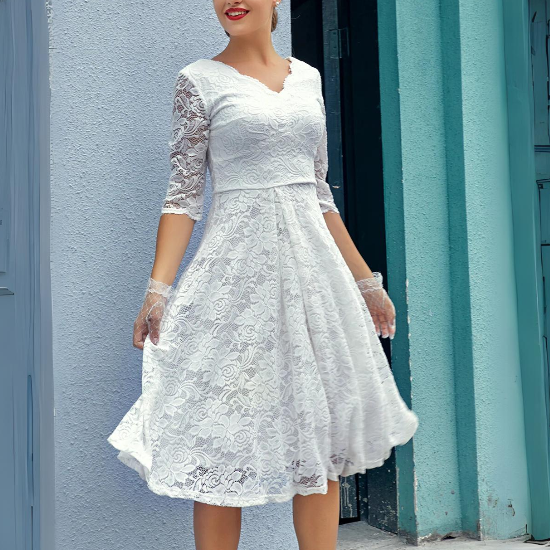 Frau in weißem Spitzenkleid mit Dreiviertelärmeln vor blauer Wand.