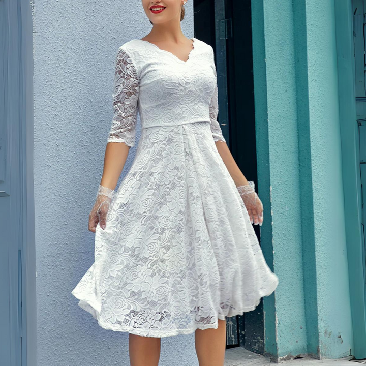 Frau in weißem Spitzenkleid mit Dreiviertelärmeln vor blauer Wand.