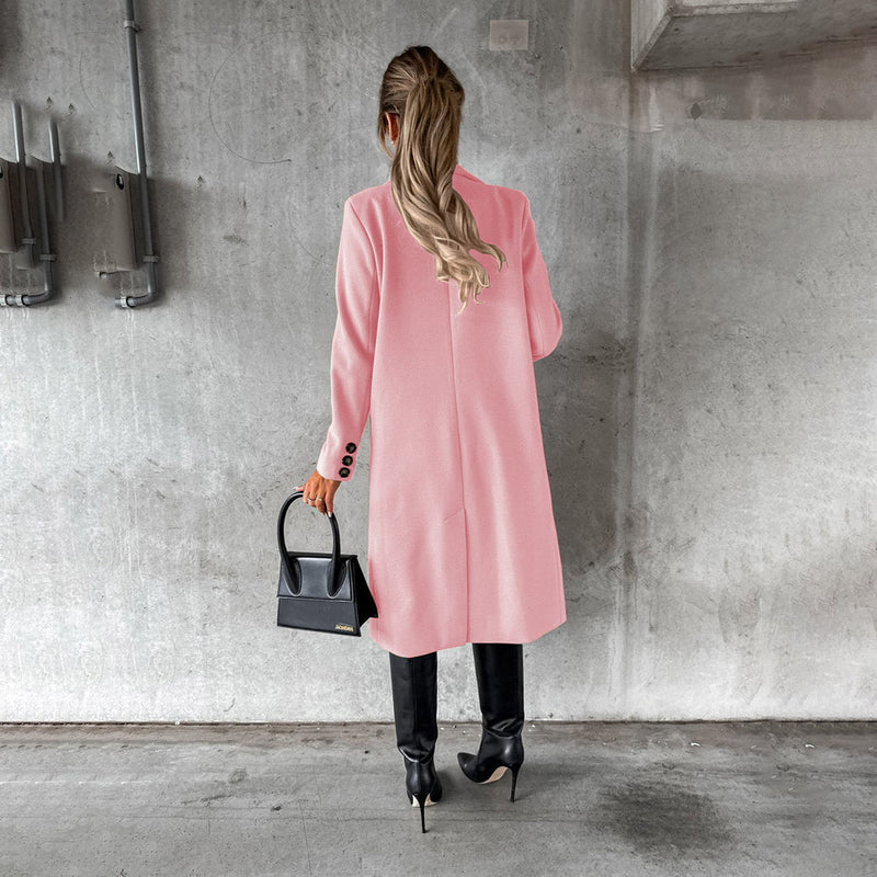 Frau in rosa Wintermantel, schwarze Stiefel, hält schwarze Handtasche, Betonhintergrund.