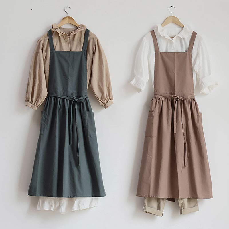 Zwei Schürzenkleider aus Baumwolle, beige und grau, mit Taschen, auf Kleiderbügeln.