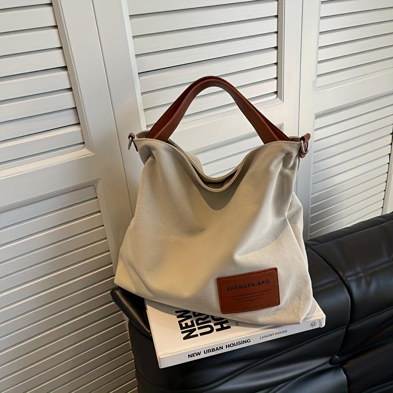 Beige Canvas-Schultertasche mit braunen Ledergriffen auf schwarzem Sofa, modisch und geräumig.