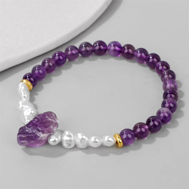 Amethyst-Armband mit weißen Perlen, lila Edelsteinen und goldenen Akzenten auf grauem Hintergrund.