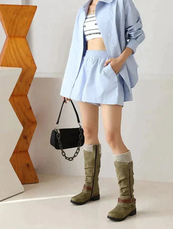 Frau in blauer Bluse und Rock, trägt olivgrüne Stiefel, hält schwarze Handtasche.