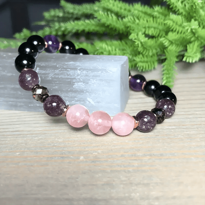 Armband aus Rosenquarz und Amethyst, handgefertigt, auf Holztisch mit grünen Pflanzen im Hintergrund.