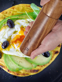 Hand mahlt Pfeffer über Avocado-Ei-Fladenbrot mit Oliven, Holz-Pfeffermühle, Frühstücksgericht.