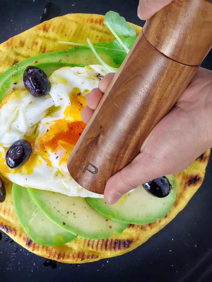 Hand mahlt Pfeffer über Avocado-Ei-Fladenbrot mit Oliven, Holz-Pfeffermühle, Frühstücksgericht.