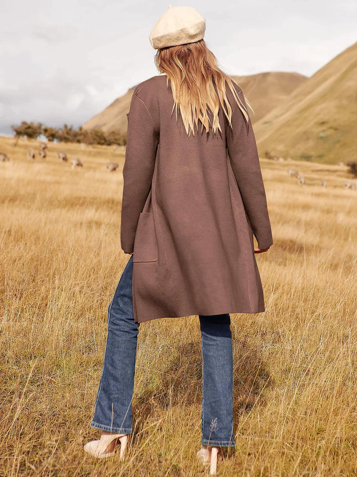 Frau in braunem Mantel und Jeans auf Wiese, beige Mütze, Herbstmode, Naturhintergrund.