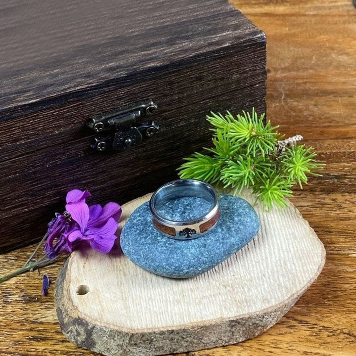 Silberring mit Baumgravur auf Holzscheibe, lila Blume, Holzkiste im Hintergrund.